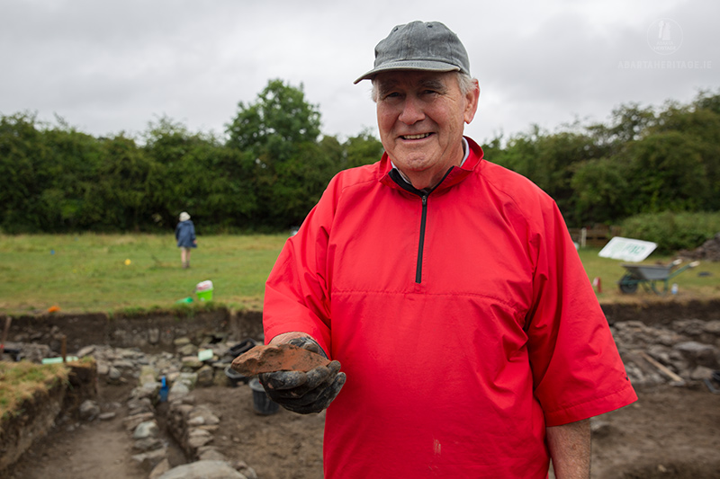 Anthony Neville in a bright red coat holding a piece of medieval pottery that he found during the excavation at Beaubec